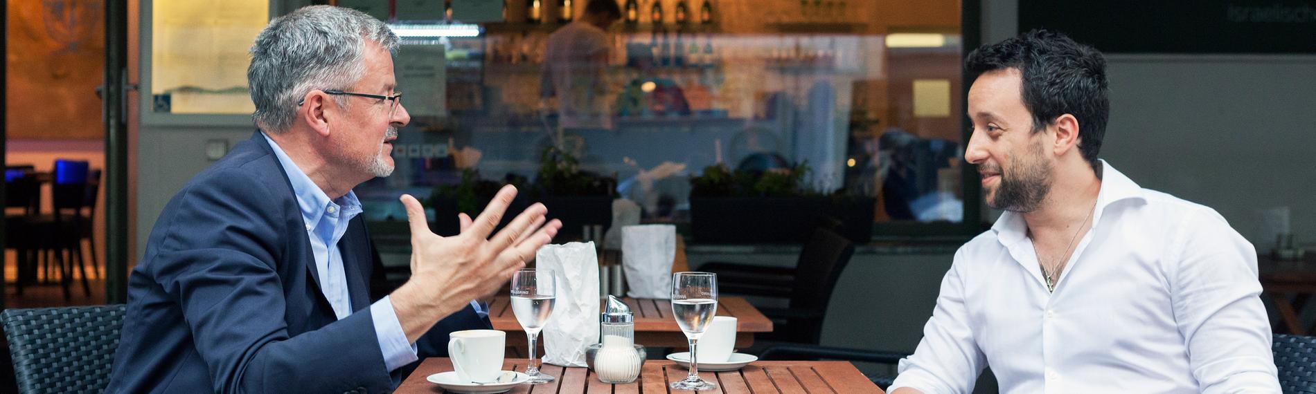 Christopher Clark und Yorai Feinberg sitzen sich an einem Café-Tisch im Freien gegenüber und diskutieren.