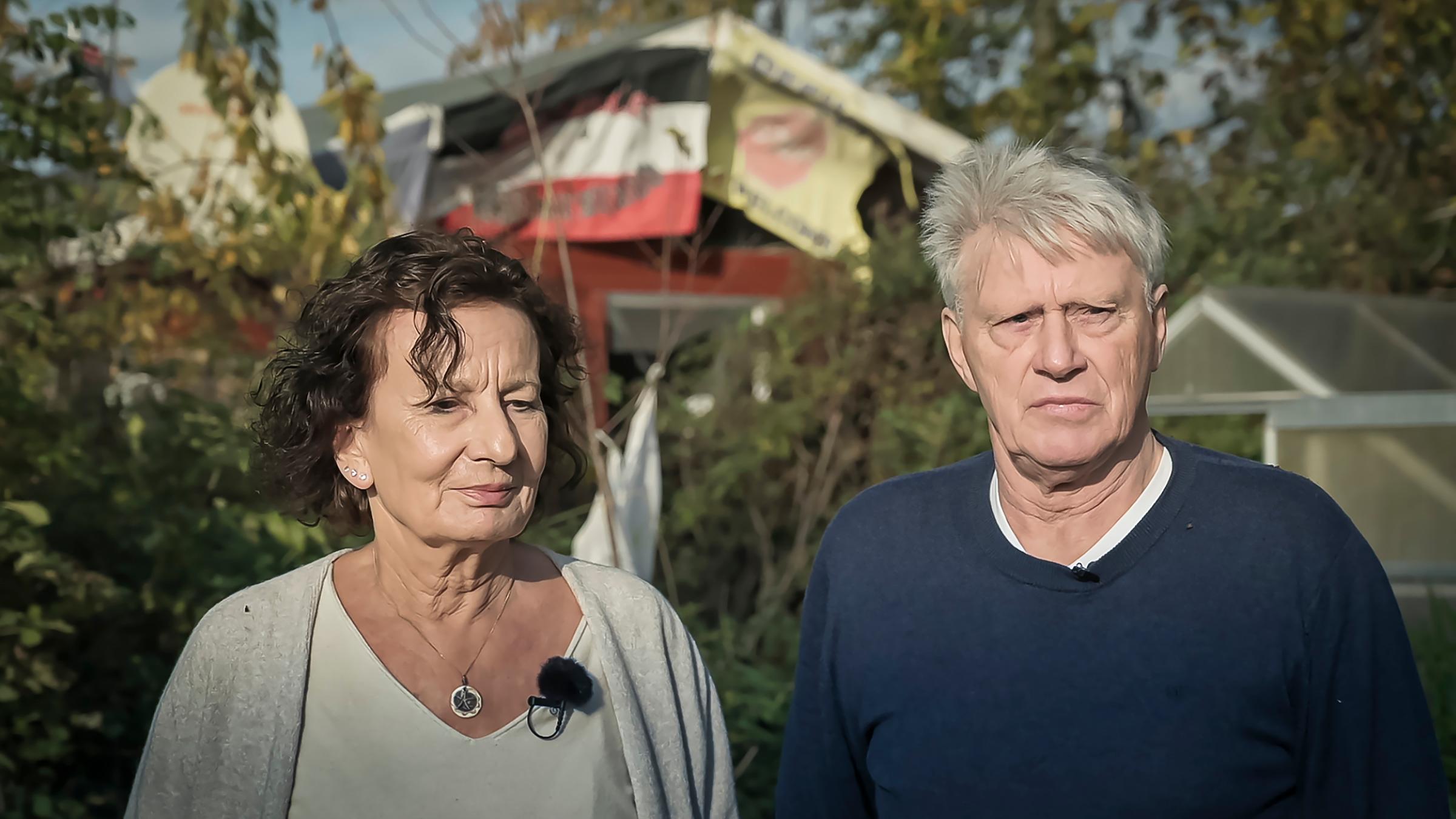 "37°: Mein Nachbar, der Neonazi - Bleiben oder wegziehen?": Sylvia und Armin Joos stehen in ihrem Garten in Hörstgen (Nordrhein-Westfalen). Sylvia hat gelocktes kinnlanges Haar und trägt ein weißes T-Shirt, darüber eine beige Strickjacke. Armin hat graue Haare und trägt einen blauen Pulli. Im Hintergrund hängt ein Banner des Nachbarn mit den Aufschriften "Nationaler Widerstand" und "Refugees Not Welcome" sowie eine Schwarze Sonne. Diese Banner hängen an der Grenze zu Joos’ Garten und sind nur vom Ehepaar einsehbar.