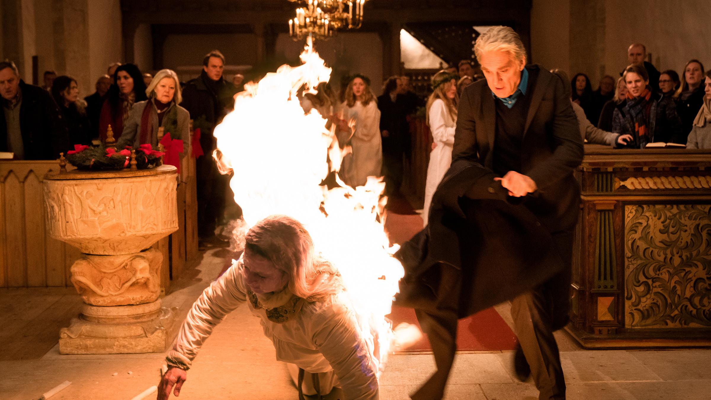  "Der Kommissar und das Meer - Lichterfest": Während der feierlichen Zeremonie brennt Malin Forstberg (Ella Lundström) ihr Umhang sie kniet auf dem Boden. Robert Anders (Walter Sittler) eilt sofort zu Hilfe und versucht mit seinem Mantel die Flammen zu löschen. Im Hintergrund schauen die Menschen in den Bankreihen entsetzt vor zu Malin.