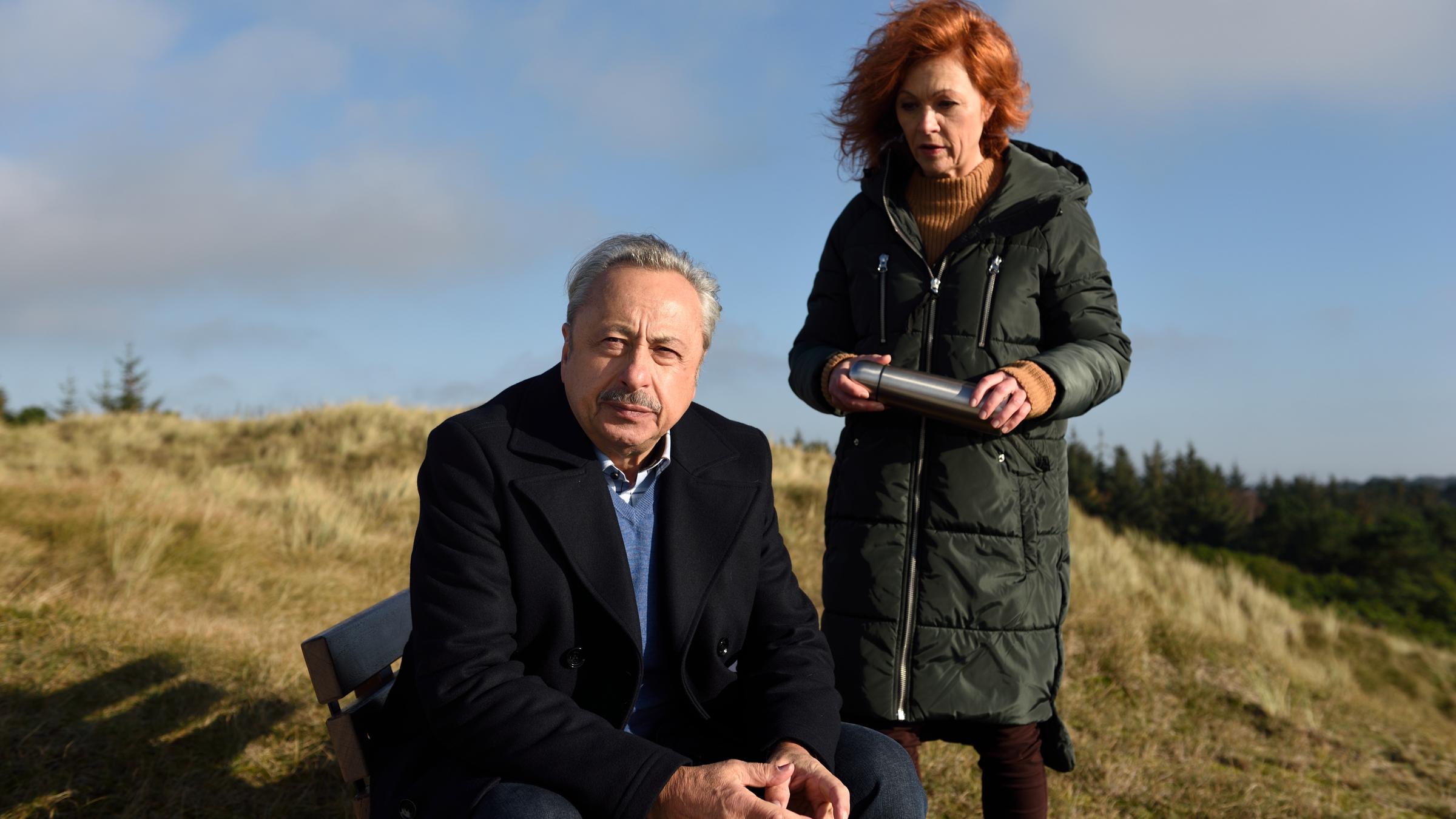 "Stubbe - Tod auf der Insel": In den begrünten Dünen steht Marlene hinter Stubbe, der auf einer Bank sitzt und in die Ferne schaut, und redet auf ihn ein.