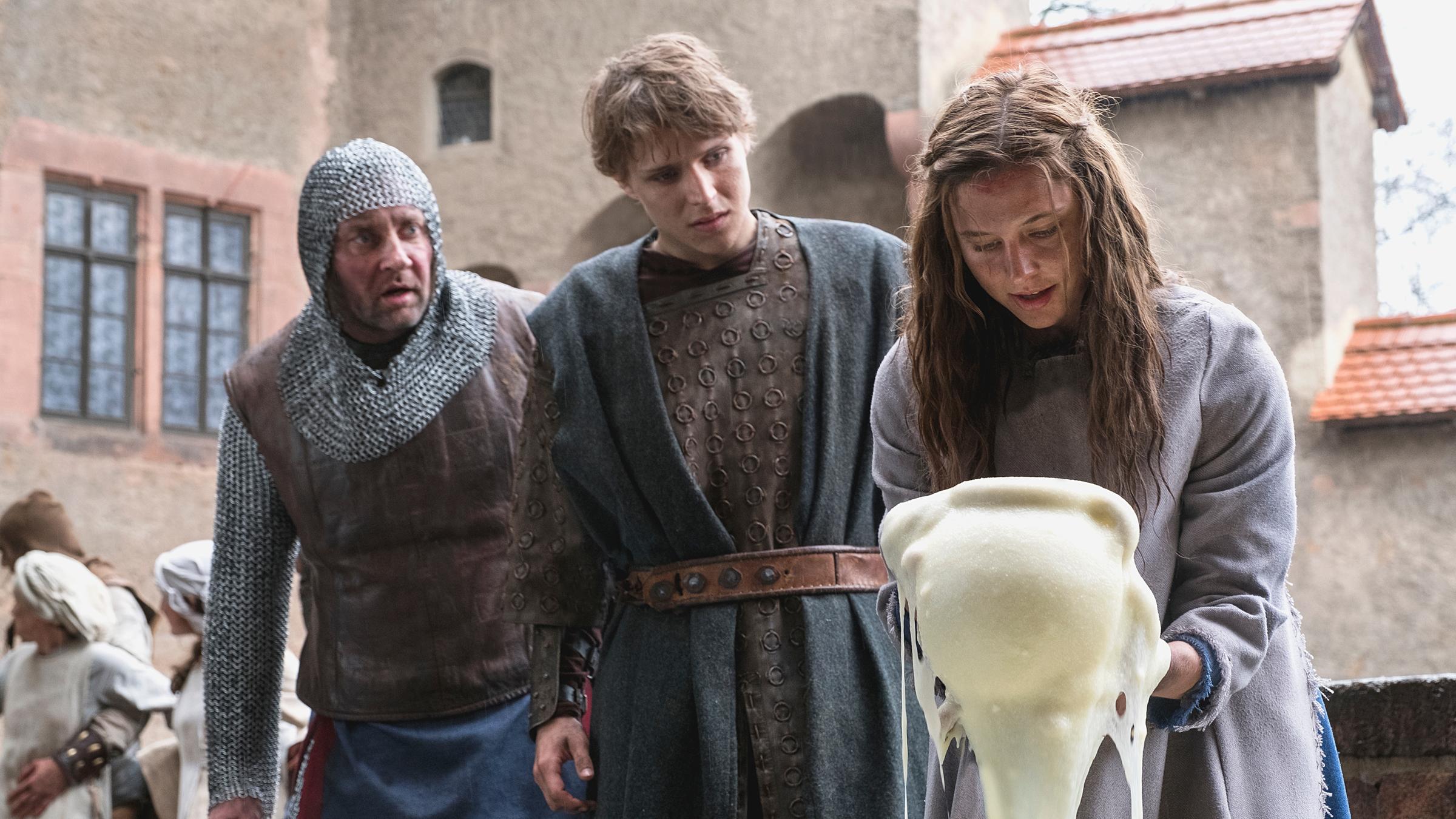 "Der süße Brei": Vor dem Hintergrund eines Innenhofes einer Burg stehen Hauptmann (Marko Dyrlich), Veit (Merlin Rose) und Jolanthe (Svenja Jung) nebeneinander. Der Hauptmann trägt ein mittelalterliches Kettenhemd, der etwa 25-jährige Veit trägt einen knielangen, ledernen Rock mit wollenem Überwurf. Beide Männer schauen stauend auf Jola. Das in einen aschgrauen Überwurf gekleidete junge Mädchen hält einen Topf in der Hand, aus dem ein Strom von süßem Brei auf den Boden fließt.