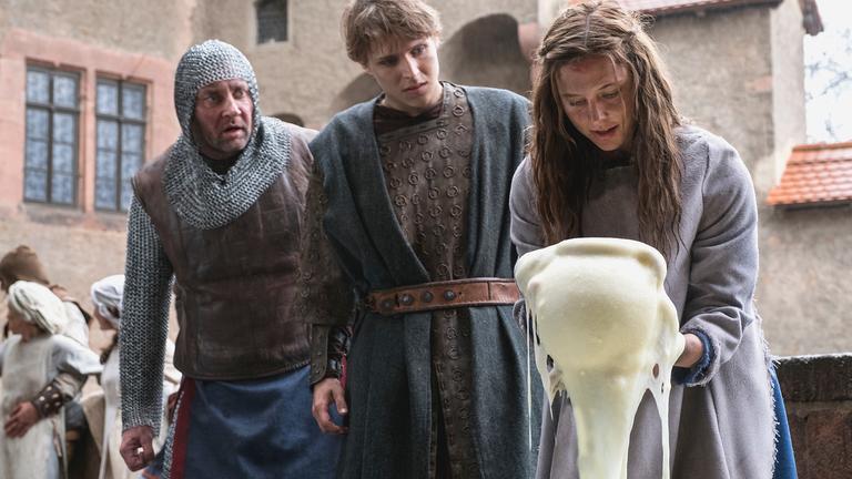 "Der süße Brei": Vor dem Hintergrund eines Innenhofes einer Burg stehen Hauptmann (Marko Dyrlich), Veit (Merlin Rose) und Jolanthe (Svenja Jung) nebeneinander. Der Hauptmann trägt ein mittelalterliches Kettenhemd, der etwa 25-jährige Veit trägt einen knielangen, ledernen Rock mit wollenem Überwurf. Beide Männer schauen stauend auf Jola. Das in einen aschgrauen Überwurf gekleidete junge Mädchen hält einen Topf in der Hand, aus dem ein Strom von süßem Brei auf den Boden fließt.