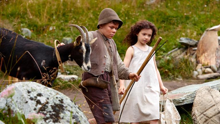 "Heidi": Heidi (Anuk Steffen) und Peter (Quirin Agrippi) laufen gemeinsam über die Almwiese, zwischen Steinen und Felsbrocken entlang. Sie werden von einer Ziege begleitet, die von Peter am Halsband geführt wird. In der anderen Hand hält er seinen Hirtenstab. Heidi trägt einen Stoffbeutel.