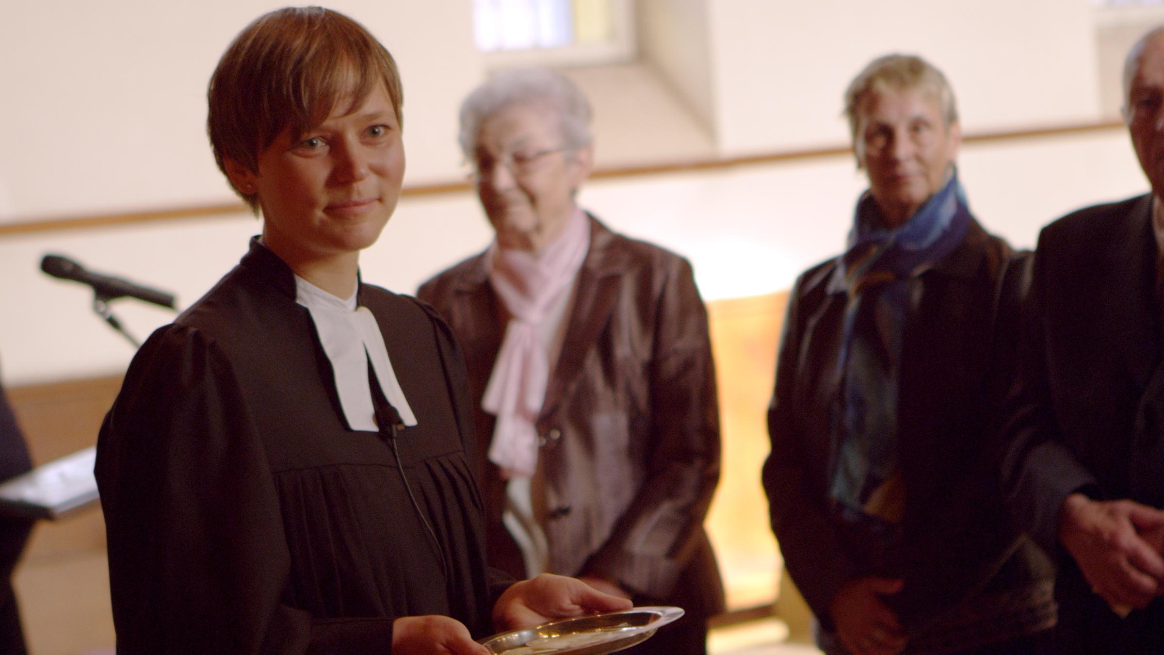 "Himmel, Herz und Hindernisse - Eine Pfarrerin packt an": Pfarrerin Irmela Büttner in ihrer Gemeinde. Sie hält eine leere Schale in den Händen, im Hintergrund sind einige ältere Menschen zu sehen.