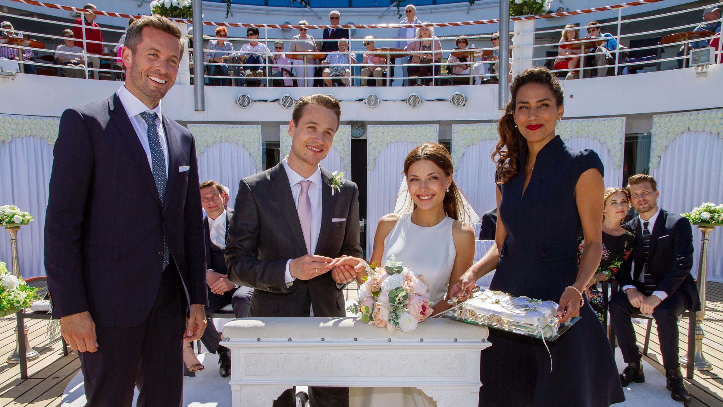 "Kreuzfahrt ins Glück - Hochzeitsreise ins Piemont": Das Brautpaar Smilla (Lena Meckel) und Carsten (Max Felder) knien auf einer weißen Kirchenbank, die an Deck des Schiffs steht. Neben ihnen stehen die Hochzeitsplaner Tom (Jan Hartmann) und Laura (Amy Mußul), die ein Tablett mi den Ringen hält. Im Hintergrund sitzen und stehen andere Gäste der Reise, unter anderen auch Oliver Schäfer (Tom Mikulla), Sabine (Tina Ruland) und Nils Ebert (Raul Richter).