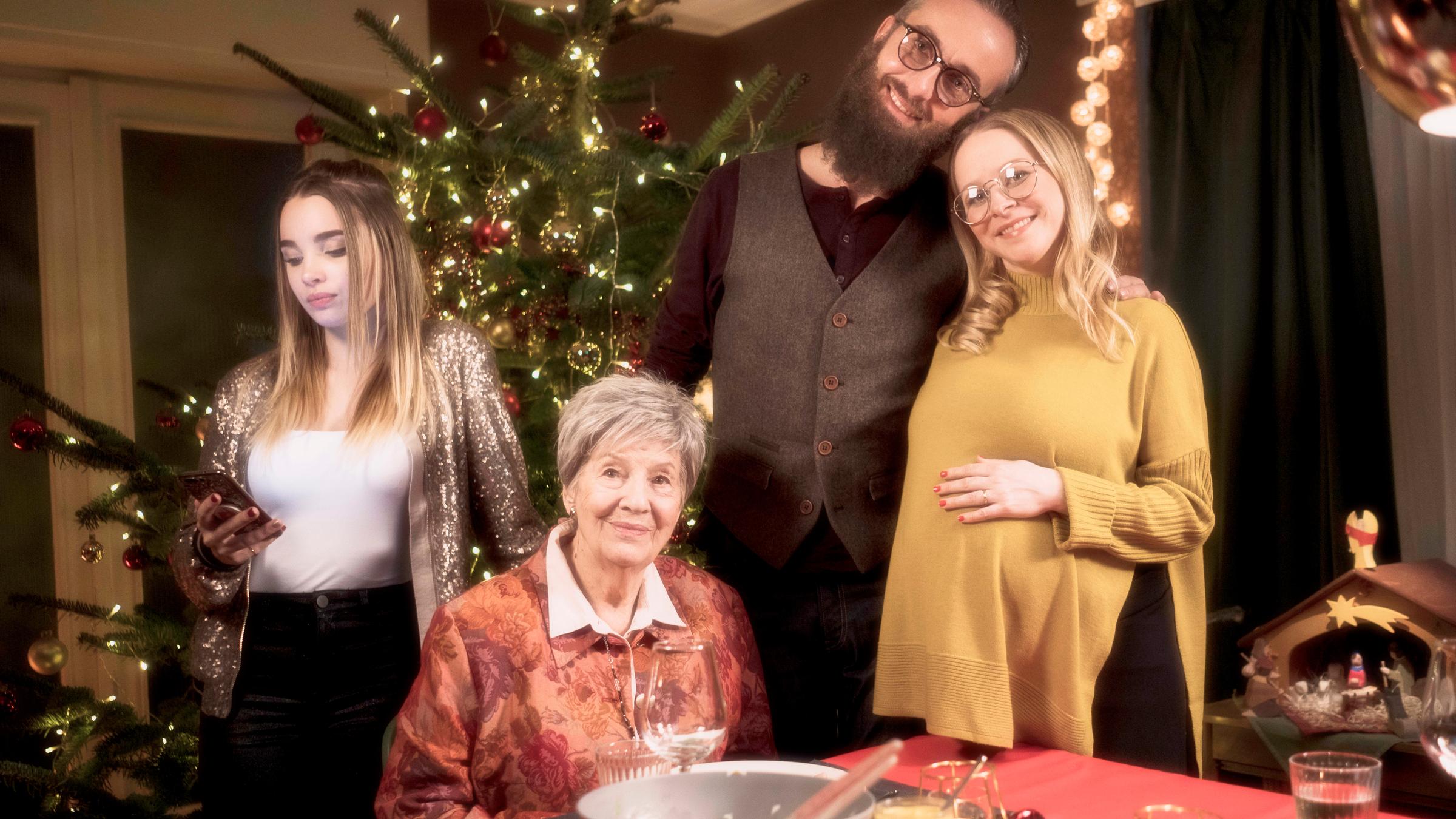 "Böhmermanns perfekte Weihnachten": Kai (Jan Böhmermann) steht mit der Schwangeren Jette (Jasmin Schwiers) im weihnachtlich geschmückten Zimmer. Nachbarin Traudel Herold (Ilse Strambowski) sitzt vor ihnen am gedeckten Esstisch. Jettes Tochter Lea (Juliette Madeleine Jozwiak) steht etwas abseits links, und schaut auf ihr Smartphone. Im Hintergrund ist ein geschmückter Weihnachtsbaum und eine Weihnachtkrippe zu sehen.
