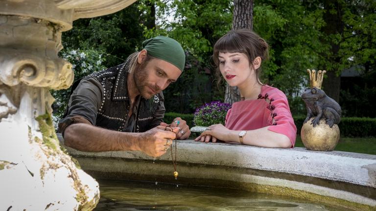 "Herzkino.Märchen: Der Froschkönig": Jimi (Bernd-Christian Althoff) und Isabelle (Anuschka Tochtermann) sitzen am Rande eines Brunnens, auf dem ein Frosch auf einer goldenen Kugel sitzt. Er hält eine Kette mit einer goldenen Kugel in der Hand.