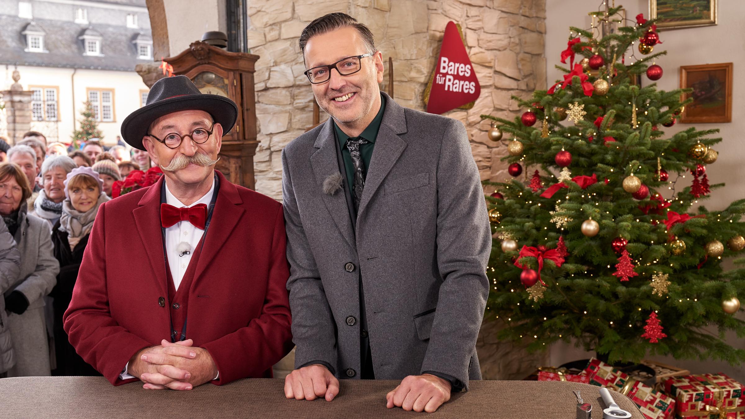 "Bares für Rares - Deutschlands größte Trödel-Show": Moderator Horst Lichter und Experte Detlev Kümmel stehen hinter einem Tisch und lächeln in die Kamera. Hinter ihnen ist ein Weihnachtsbaum und Publikum zu sehen.