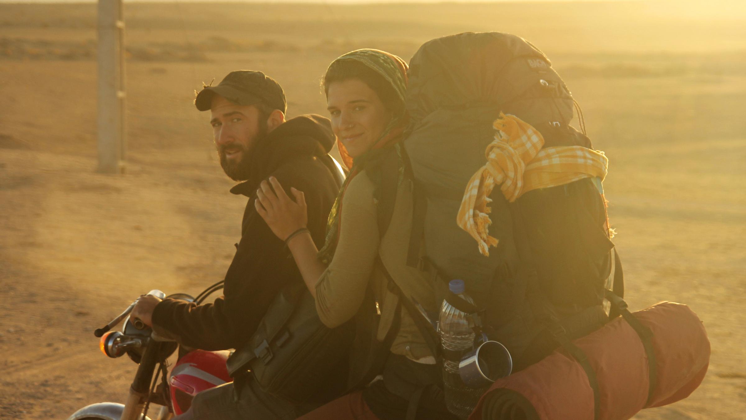 „Weit“: Ein junger Mann mit Bart und Baseballkappe sitzt am Lenker eines Motorrads und schaut in die Kamera. Hinter ihm sitzt eine junge Frau, die sich an seiner Schulter festhält, die Haare mit einem Tuch bedeckt hat und einen vollbepackten Rucksack auf den Schultern trägt. Im Hintergrund ist eine weite Steppenlandschaft unscharf und in einem goldgelben Licht der Abendsonne zu sehen.