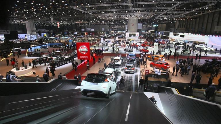 "Ausgebremst - Überlebenskampf der Autobauer": Blick von oben in eine große Messehalle mit vielen ausgestellten Autos und Menschen. Im Vordergrund ist das Firmenlogo von Nissan zu erkennen, das von der Decke herunterhängt.