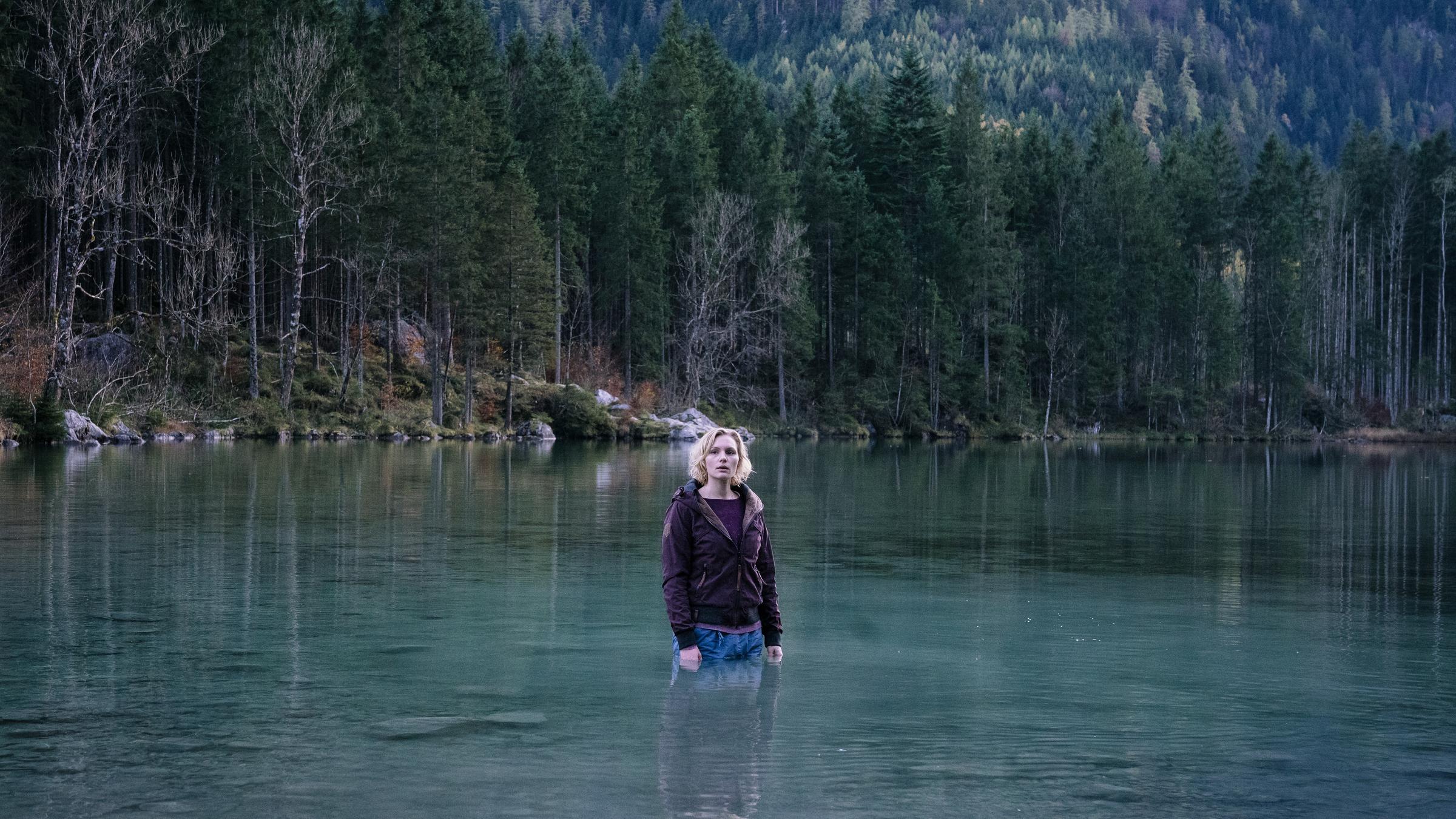 "Rufmord": Die vollständig bekleidete Luisa (Rosalie Thomass) steht bis zu den Hüften in einem See. Im Hintergrund ragen Bäume am steilen und felsigen Ufer des grün schimmernden Sees in die Höhe.