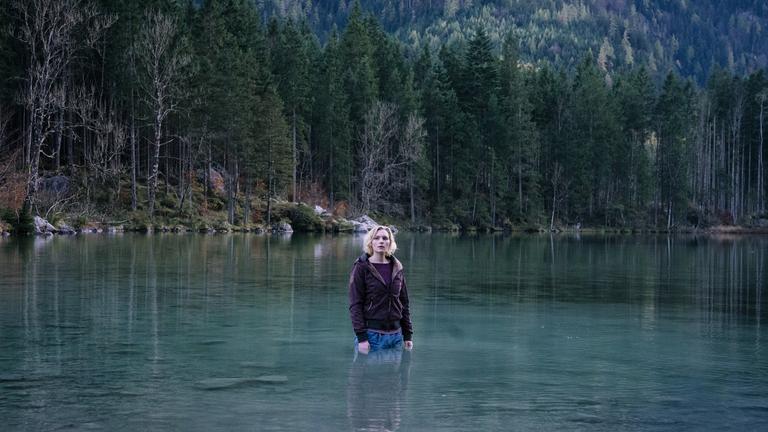 "Rufmord": Die vollständig bekleidete Luisa (Rosalie Thomass) steht bis zu den Hüften in einem See. Im Hintergrund ragen Bäume am steilen und felsigen Ufer des grün schimmernden Sees in die Höhe.
