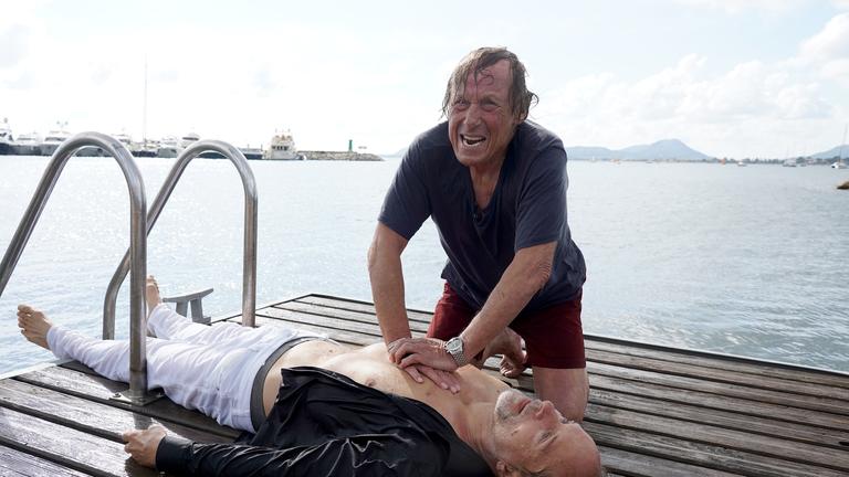 "Matula - Tod auf Mallorca": Matula (Claus Theo Gärtner) kniet durchnässt auf einem Steg am Meer und macht macht eine Herzdruckmassage bei dem leblosen Ingo Schumann (Jochen Horst), den er offensichtlich gerade aus dem Wasser gezogen hat. Im Hintergrund sieht man einige Boote im Wasser.
