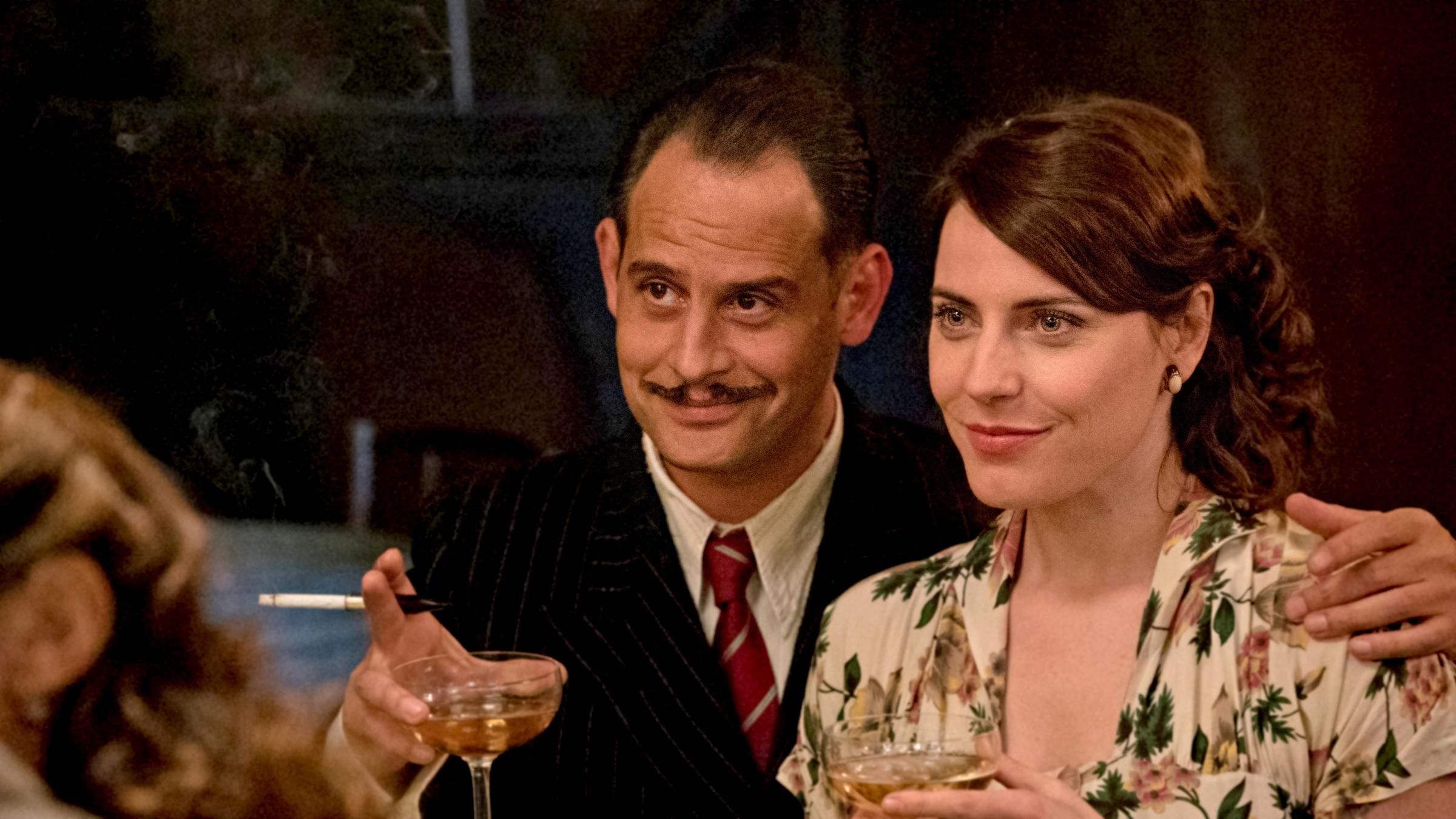 "Es war einmal in Deutschland": David (Moritz Bleibtreu) hat den Arm um Saras (Antje Traue) Schulter gelegt. Er hält eine Zigarette und ein Glas Champagner in der Hand, beide lächeln.
