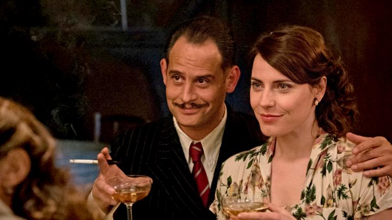 "Es war einmal in Deutschland": David (Moritz Bleibtreu) hat den Arm um Saras (Antje Traue) Schulter gelegt. Er hält eine Zigarette und ein Glas Champagner in der Hand, beide lächeln.