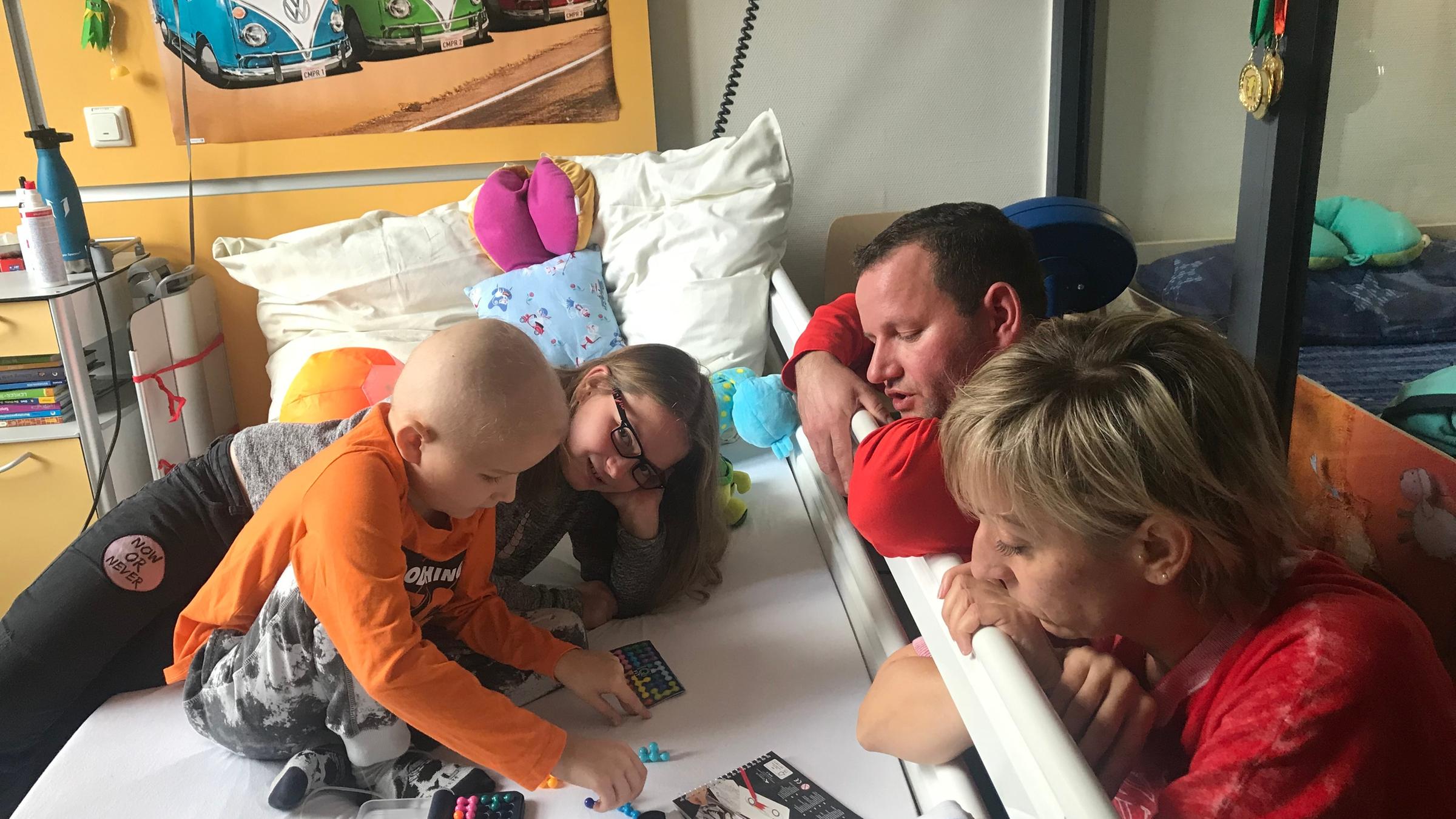 "37° Keine Zeit für Tränen - Familien mit krebskranken Kindern": Fabio spielt auf seinem Bett im Krankenhaus. Seine Schwester ist seitlich hinter ihm. Vater und Mutter schauen von der anderen Seite zu.