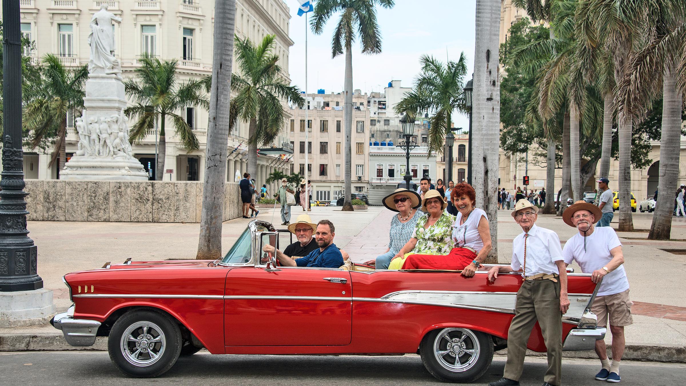 "Mit 80 Jahren um die Welt": Theo, Steven Gätjen, Gisela, Ruth und Marianne sitzen in einem roten Oldtimer-Cabrio, Ernst und Nauke lehnen stehen am Heck. Im Hintergrund Palmen und Gebäude des "Parque Central". Alle schauen freundlich in die Kamera.