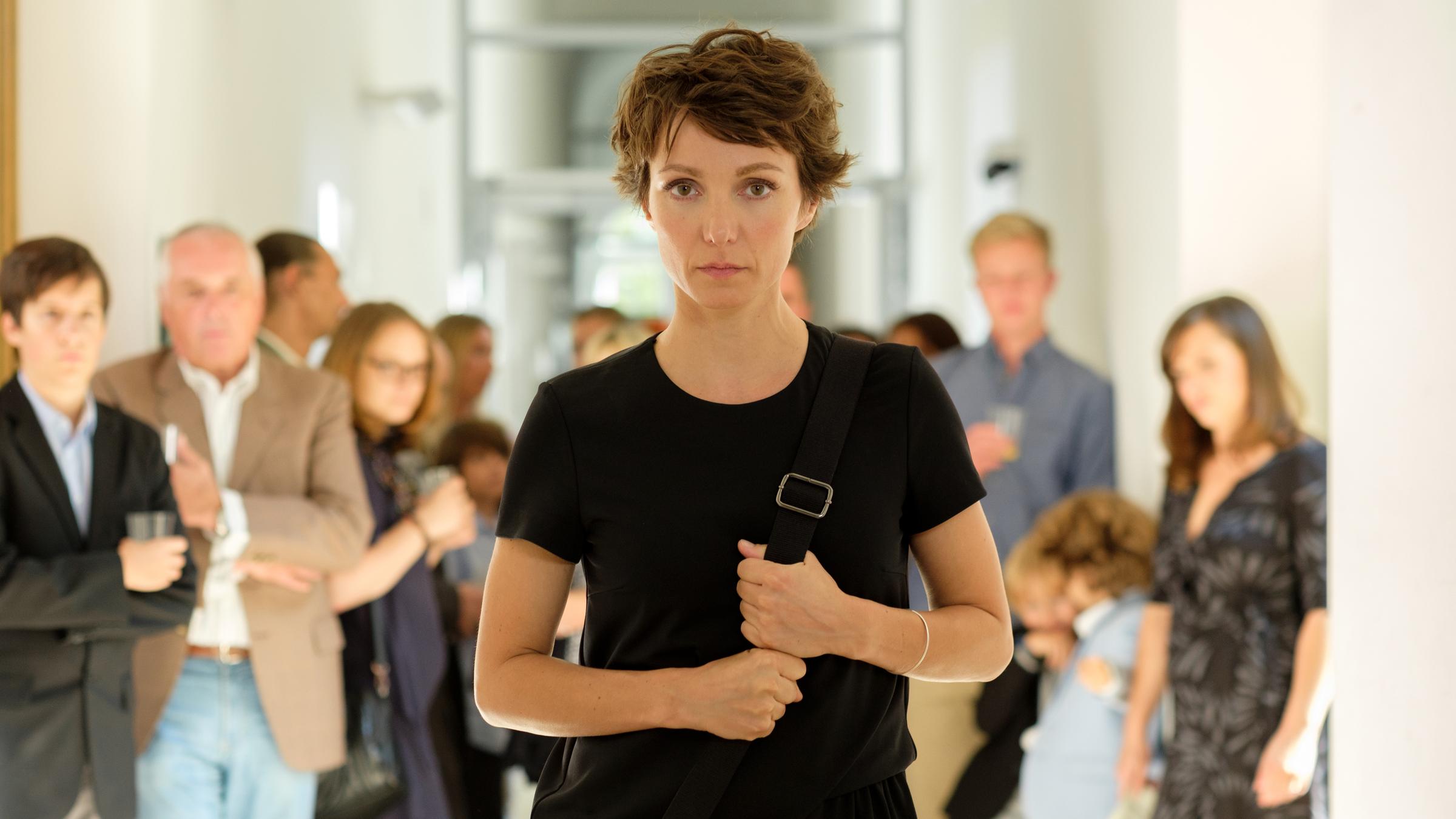 "Balanceakt": Marie blickt forsch und entschlossen direkt in die Kamera. Sie trägt ein schwarzes Sommerkleid und hat eine Umhängetasche über der Schulter, deren Riemen sie mit beiden Händen packt. Im Hintergrund in der Unschärfe stehen Männer und Frauen und blicken ihr nach.