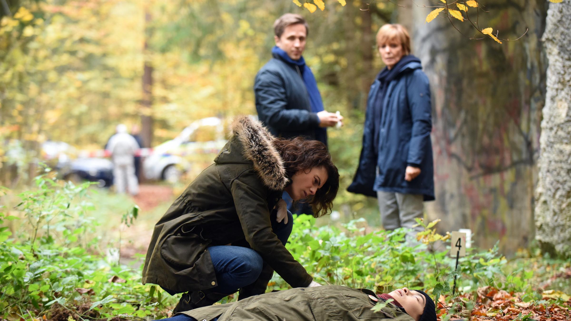 "Kommissarin Lucas - Tote Erde": Im Wald liegt eine Leiche auf dem Boden, Judith Marlow (Jördis Richter) sitzt seitlich in der Hocke daneben und untersucht sie. Im Hintergrund stehen Ellen Lucas (Ulrike Kriener) und Tom Brauer (Lasse Myhr).