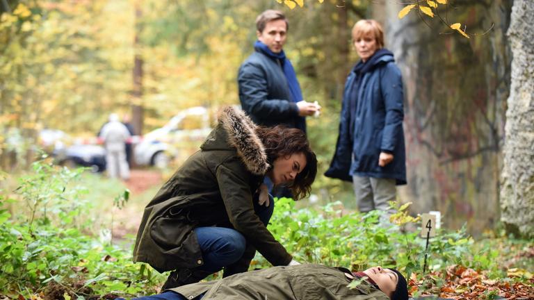 "Kommissarin Lucas - Tote Erde": Im Wald liegt eine Leiche auf dem Boden, Judith Marlow (Jördis Richter) sitzt seitlich in der Hocke daneben und untersucht sie. Im Hintergrund stehen Ellen Lucas (Ulrike Kriener) und Tom Brauer (Lasse Myhr).