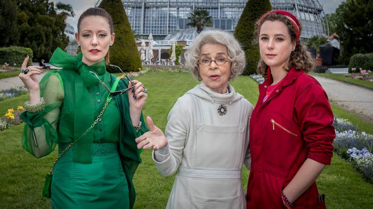 "Frau Holles Garten": Frau Holle (Cornelia Froboess) steht in der Mitte zwischen Mia Goldig (Klara Deutschmann) und ihrer Schwester Marissa (Lavinia Wilson) vor einem Gewächshaus im Botanischen Garten.