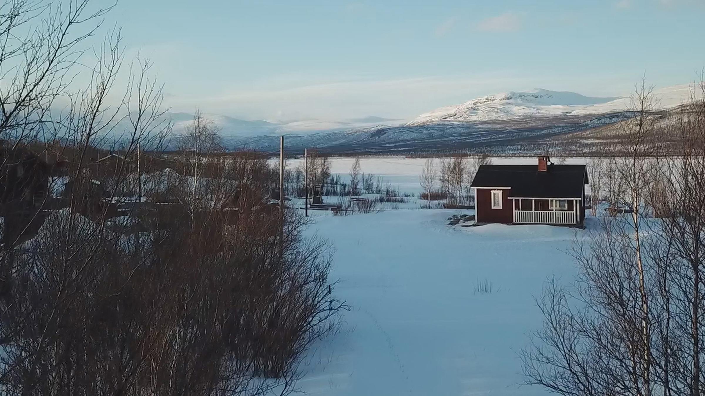 Skandinaviens versteckte Paradiese: Totale Winterlandschaft mit kahlen Sträuchern, einem Holzhaus im Mittelgrund und einem hohen Berg rechts im Hintergrund