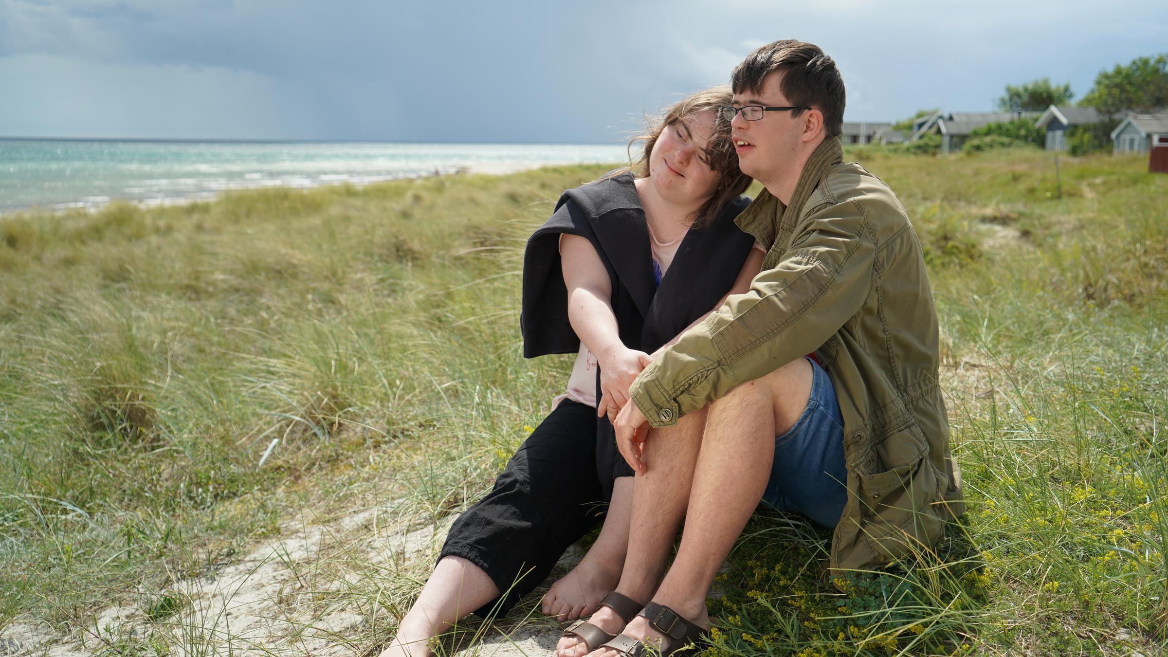 "37°: Liebe macht mutig - Zu Zweit mit Down-Syndrom": Emily und Erik, jeweils mit Down-Syndrom, sitzen auf einer Düne am Strand und lächeln verliebt.