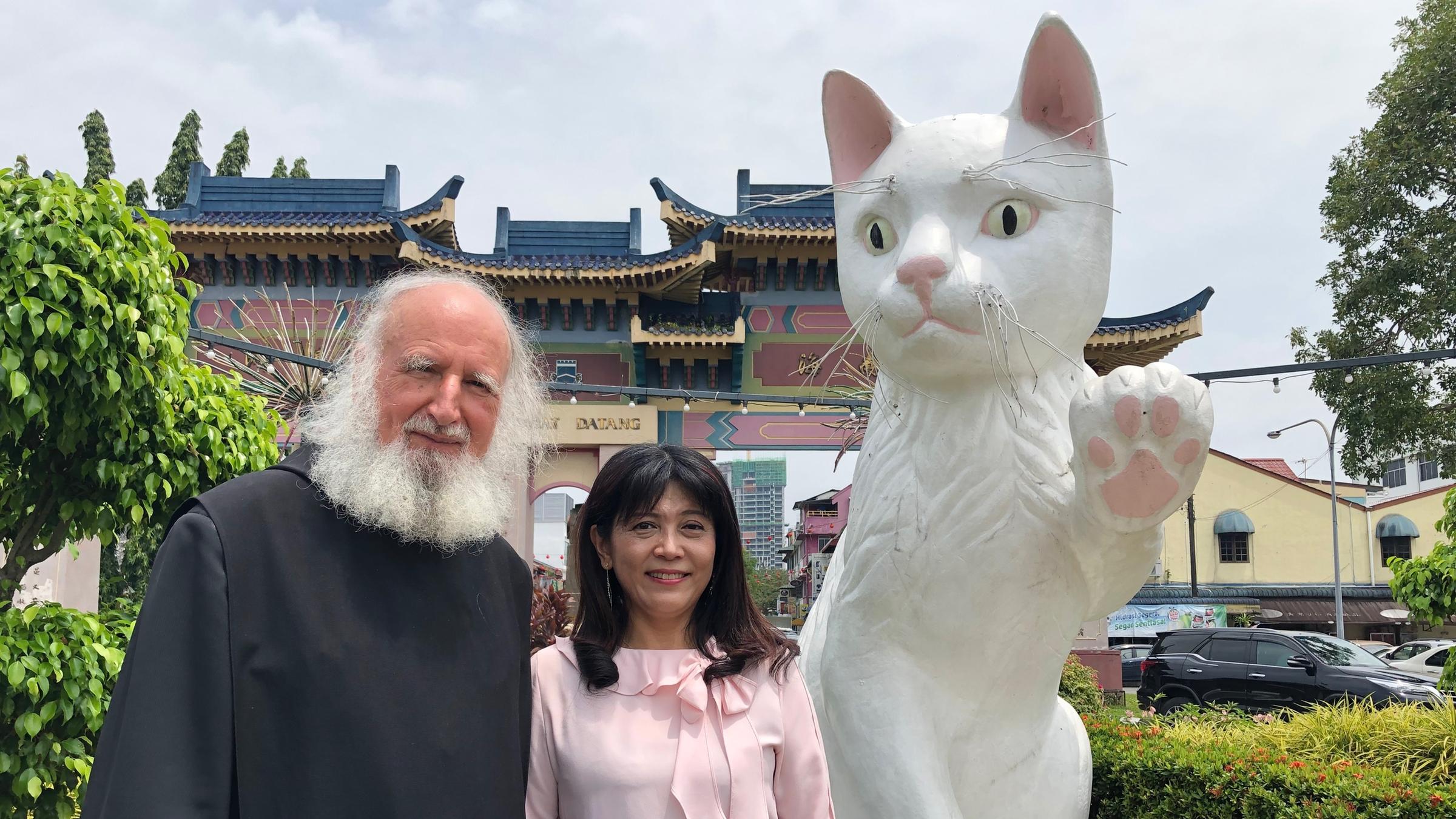 "Das Phänomen Anselm Grün": Pater Anselm Grün und Frau Wu stehen vor einem taiwanischen Gebäude.  Neben ihnen ist eine übergroße weiße Katzenskulptur.