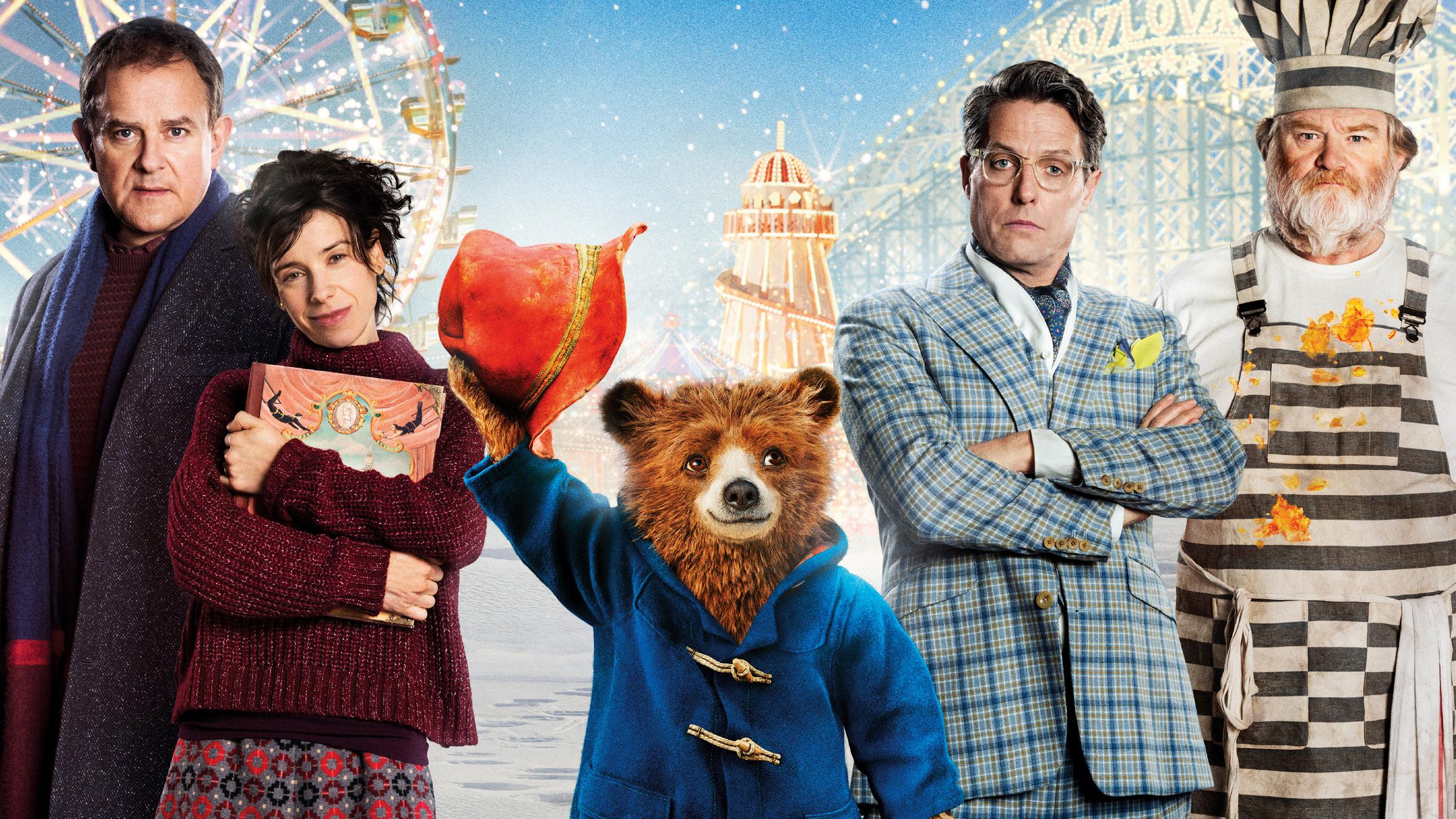 "Paddington 2": Filmplakat - Mr. und Mrs. Brown (Hugh Bonneville, Sally Hawkins), Gefängniskoch Knuckles  (Brendan Gleeson) und Schauspieler Phoenix Buchanan (Hugh Grant) stehen vor den Zeichnungen eines Riesenrads, einer Achterbahn und einem Zirkuszelt nebeneinander. Paddington hebt zum Gruß seinen Hut.