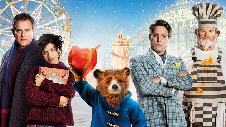 "Paddington 2": Filmplakat - Mr. und Mrs. Brown (Hugh Bonneville, Sally Hawkins), Gefängniskoch Knuckles  (Brendan Gleeson) und Schauspieler Phoenix Buchanan (Hugh Grant) stehen vor den Zeichnungen eines Riesenrads, einer Achterbahn und einem Zirkuszelt nebeneinander. Paddington hebt zum Gruß seinen Hut.