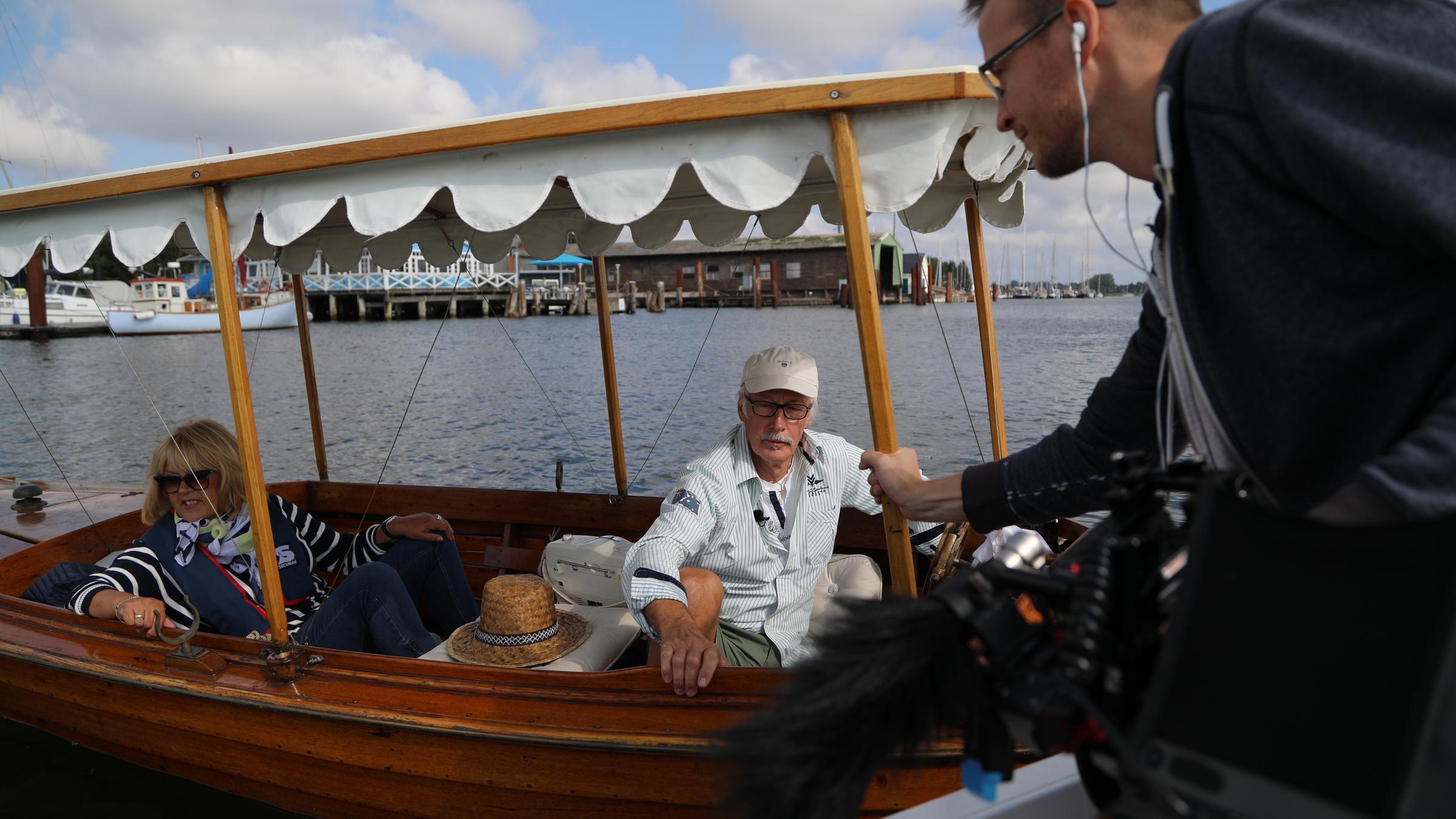 "37°: Mein letzter Tag im Betrieb": Annemarie und Hans-Werner Broderius sitzen in einem kleinen Holzboot mit Baldachin. Ein Reporter hält ihnen vom Ufer aus ein Mikrofon entgegen.