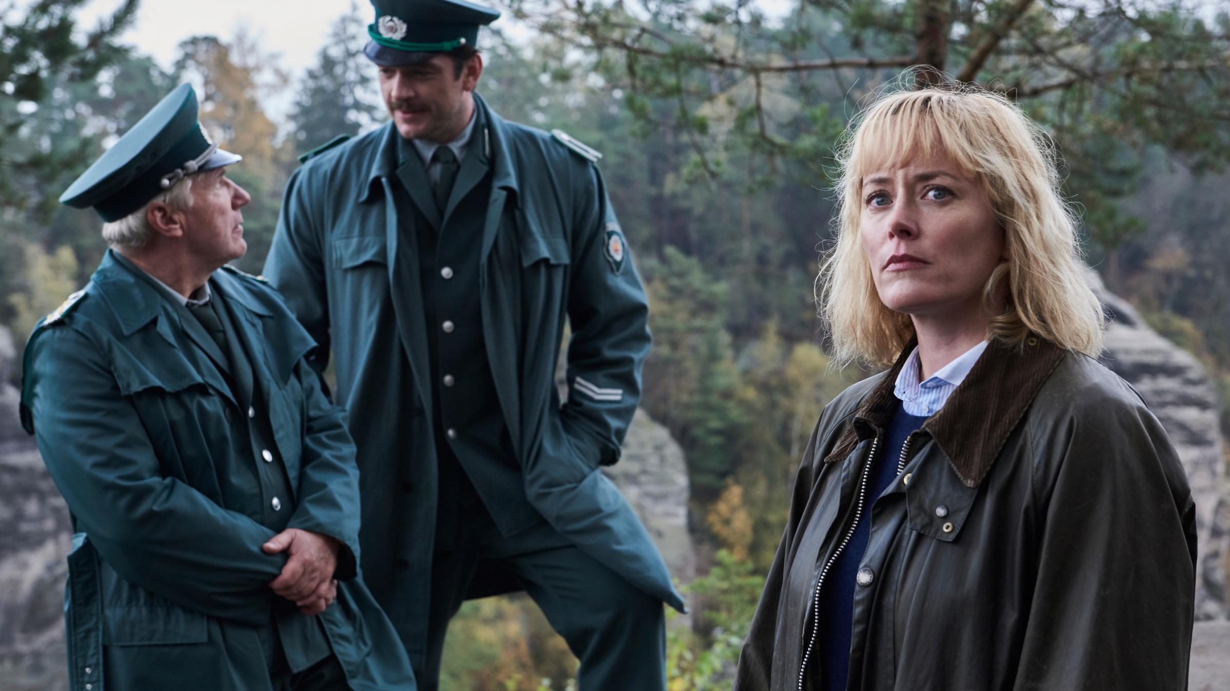 „Walpurgisnacht - Die Mädchen und der Tod (1)“: Lothar Wieditz (Jörg Schüttauf) und Karl Albers (Ronald Zehrfeld), die beide eine grüne Polizeiuniform tragen, stehen nebeneinander im Freien und schauen sich an. Leicht versetzt vor ihnen steht Nadja Paulitz (Silke Bodenbender). Im Hintergrund sind Bäume zu erkennen.