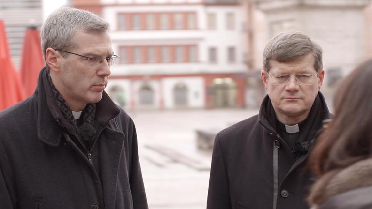 "ZDFzoom: Abschottung oder Aufbruch? - Die katholische Kirche und die Missbrauchskrise": Die Bischöfe Heiner Wilmer und Stephan Burger stehen auf einem Marktplatz im Gespräch mit dem ZDF-Team.