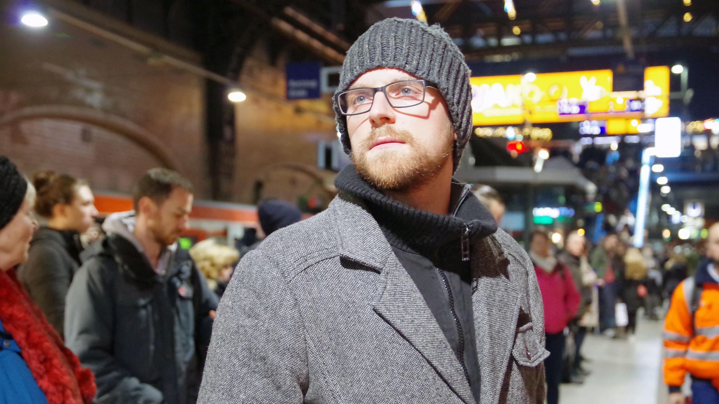 "plan b: Gemeinsam statt einsam - Vom heilsamen Miteinander": Christian Adis steht mit Mütze und Mantel bekleidet inmitten einer Menschenmenge am Bahnhof und schaut sich um.