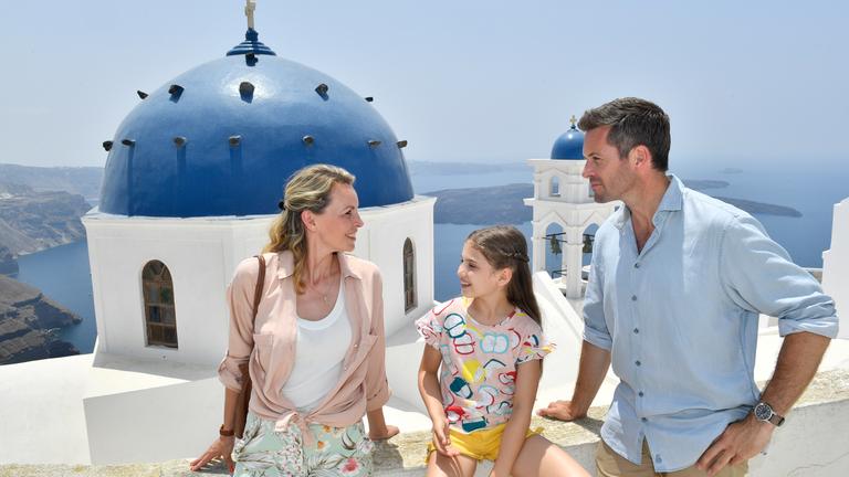 "Kreuzfahrt ins Glück: Hochzeitsreise auf die Kykladen": Zusammen mit Hochzeitsplaner Tom (Jan Hartmann) sitzen Natalie (Simone Hanselmann) und Maja (Helene Pieske) auf einer weißen Steinmauer und unterhalten sich. Im Hintergrund sieht man typische griechische Häuser, weiße Mauern und blaue Dächer, sowie das Meer.