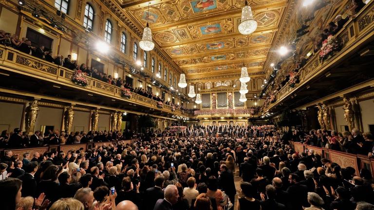 "Neujahrskonzert der Wiener Philharmoniker": Der Goldene Saal im Wiener Musikverein (hier vollbesetzt mit Publikum und Orchester auf der Bühne) ist ein prunkvoller Konzertsaal mit Logen, Balkonen,  Deckenmalerei und Kronleuchtern und anlässlich des "Neujahrskonzerts"  dekoriert mit Blumenschmuck.