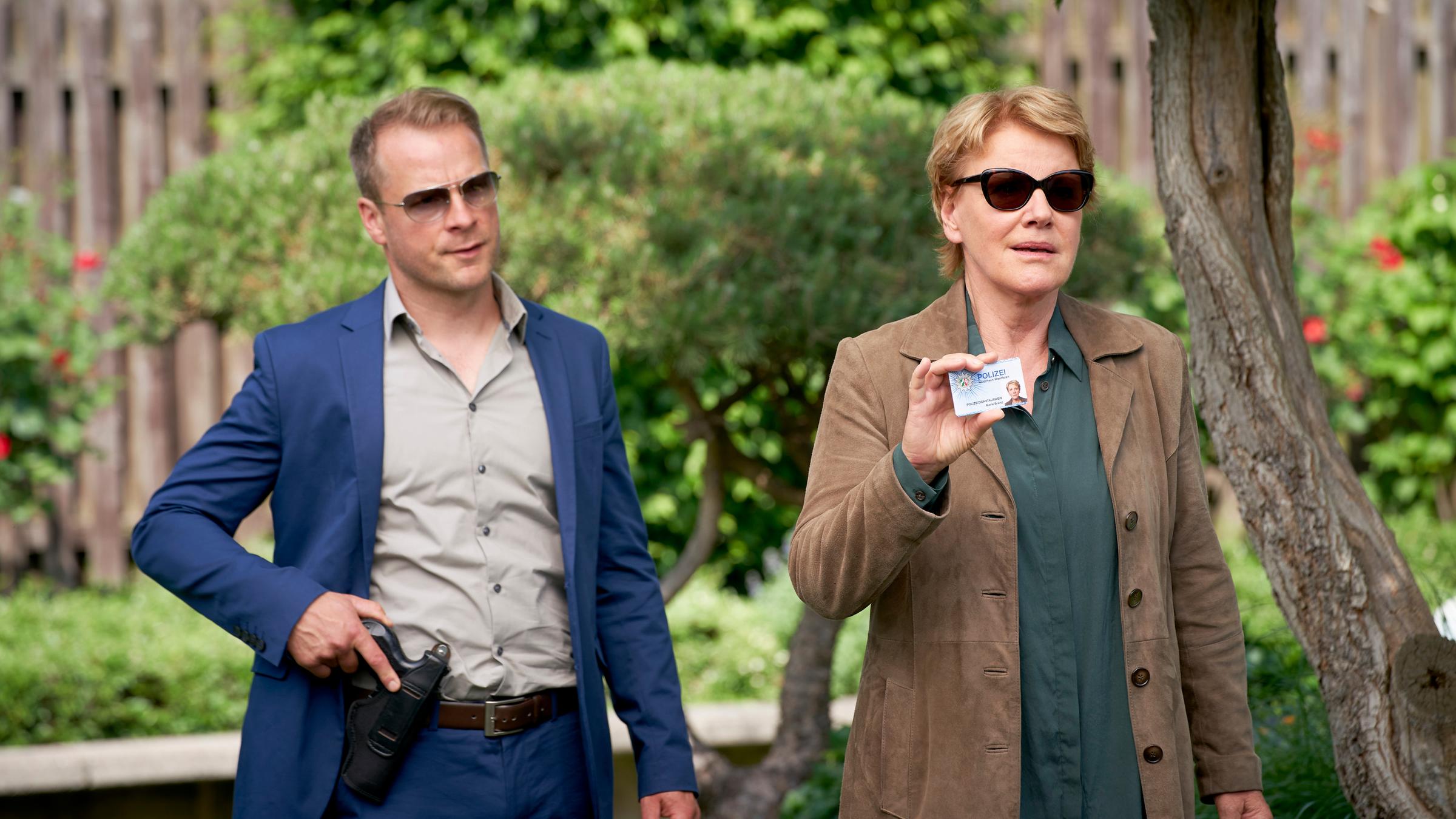 "Marie Brand und der Reiz der Gewalt": Marie Brand (Mariele Millowitsch) und Jürgen Simmel (Hinnerk Schönemann) stehen im Garten von Gregor Hoffmann. Simmel greift mit seiner rechten Hand an seine Pistole, die an seinem Gürtel befestigt ist. Marie Brand hält ihren Dienstausweis in der Hand und zeigt ihn vor. Beide tragen eine Sonnenbrille.