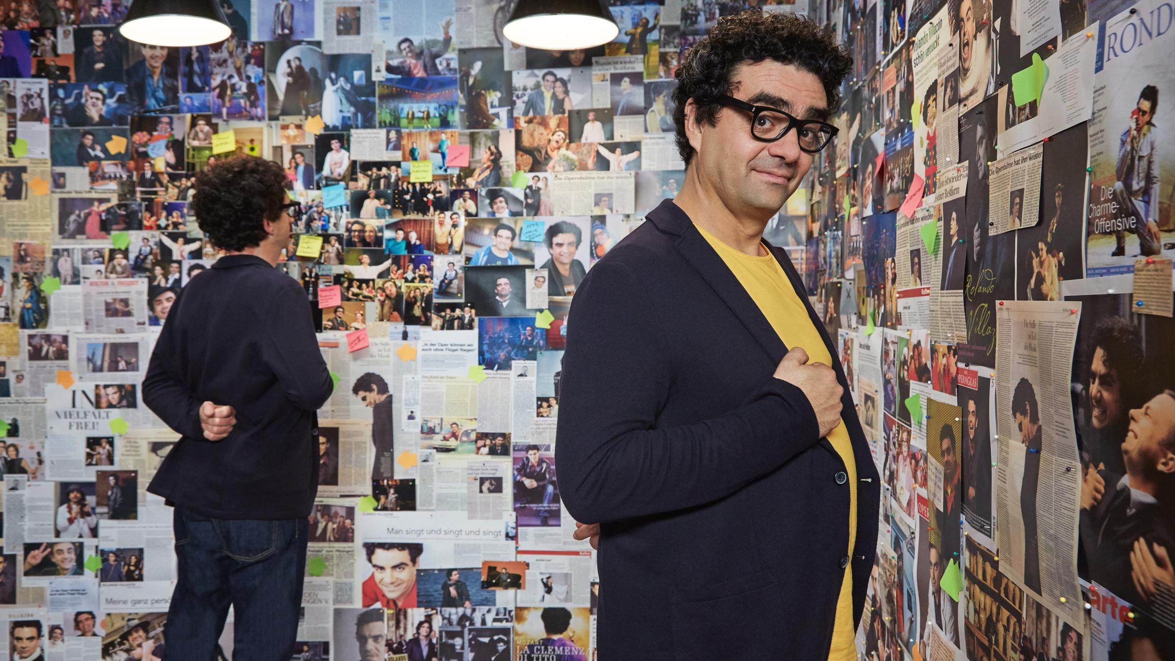 "Kessler ist... - Rolando Villazón": Rolando Villazón steht vor einer Bilderwand mit gesammelten Fotos seines bisherigen Lebens, seiner Kindheit und seiner Karriere. Er schaut verschmitzt in die Kamera und lächelt. Im Hintergrund ist, ihm den Rücken zugewandt, Michael Kessler zu sehen, der Rolando Villazón zum Verwechseln ähnlich sieht.