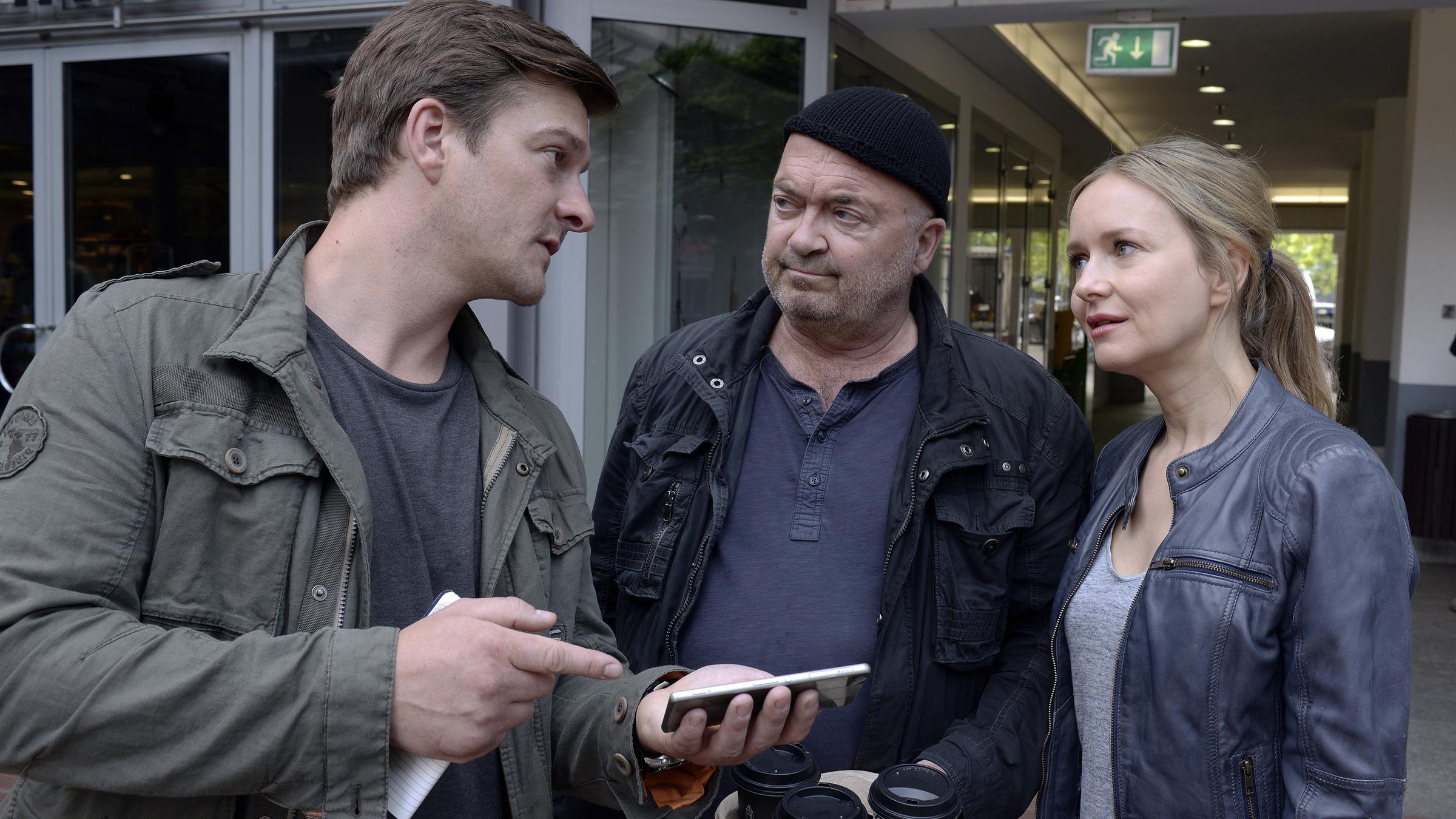 "Ein starkes Team - Eiskalt": Sebastian Klöckner (Matthi Faust), Otto Garber (Florian Martens) und Linett Wachow (Stefanie Stappenbeck) stehen vor einem Gebäude. Sebastian zeigt den beiden anderen etwas auf einem Smartphone.
