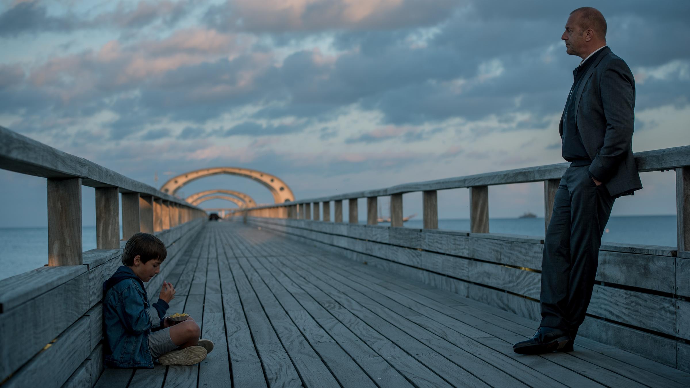 „Die verschwundene Familie (2)“: Kessler (Heino Ferch) steht auf einem Steg und lehnt sich an die Brüstung. Ihm gegenüber sitzt Tom (Timo Hack) und isst etwas.