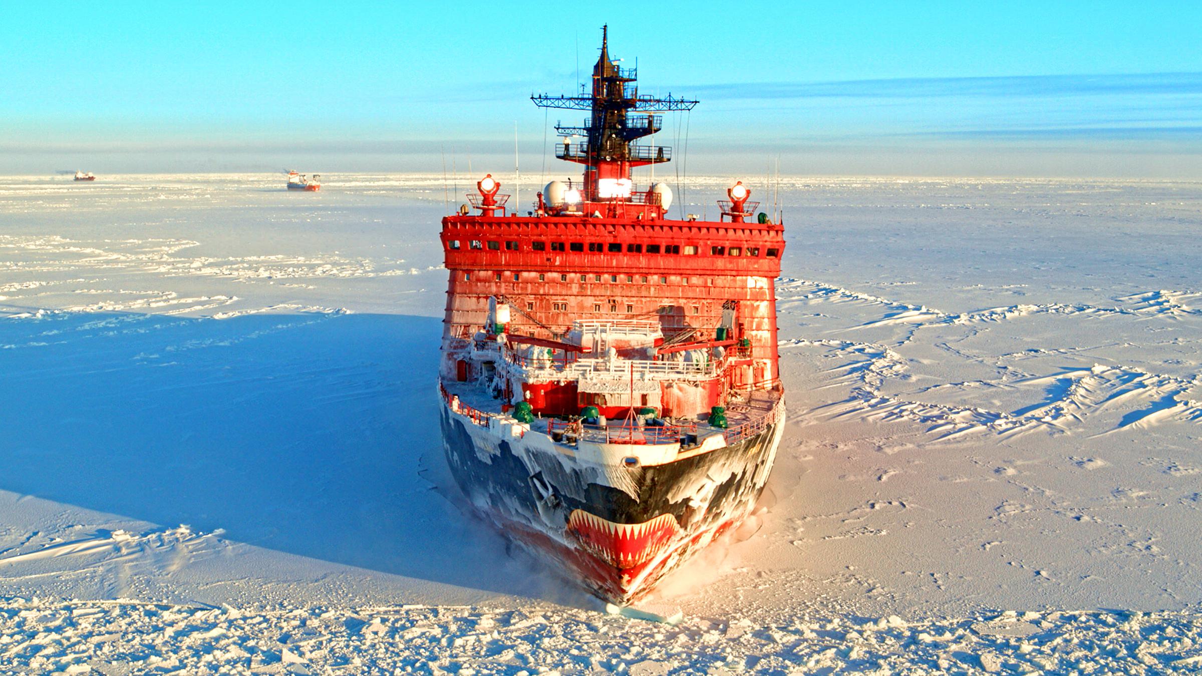 "Terra X: Russland von oben -  Nordpolarmeer und ewiges Eis": Großer, roter Eisbrecher auf einer schneebedeckten Eisfläche im Nordpolarmeer). Im Hintergrund zwei kleine Schiffe und blauer Winterhimmel.