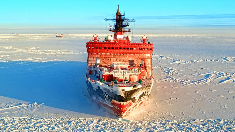 "Terra X: Russland von oben -  Nordpolarmeer und ewiges Eis": Großer, roter Eisbrecher auf einer schneebedeckten Eisfläche im Nordpolarmeer). Im Hintergrund zwei kleine Schiffe und blauer Winterhimmel.