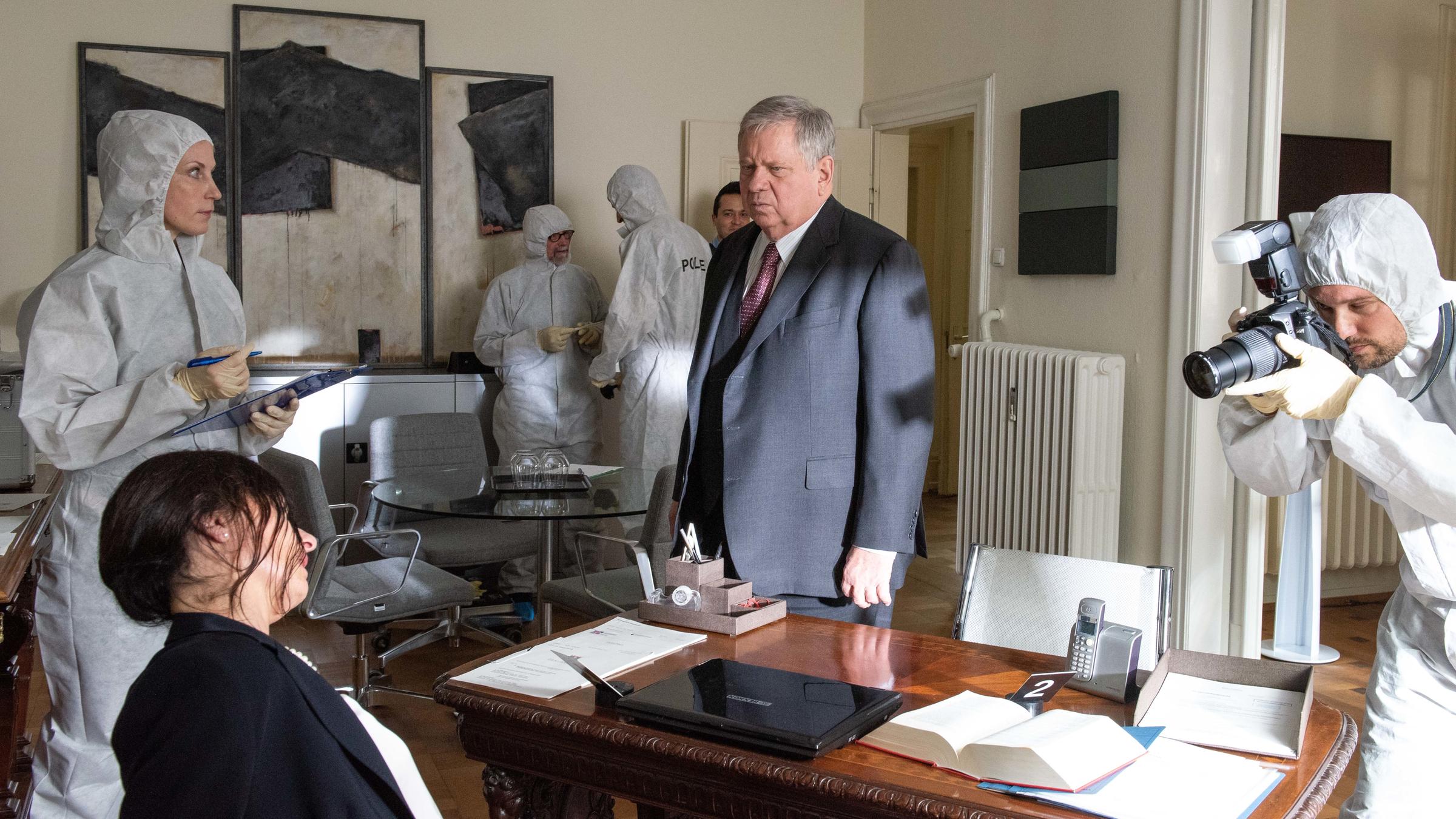 "Der Staatsanwalt - Tödlich Wohnen": Reuther (Rainer Hunold) steht in einem Arbeitszimmer und betrachtet die Leiche der getöteten Beate Schwartz (Lil von Essen), die an den Schreibtischstuhl gefesselt ist. Neben der Leiche steht Dr. Judith Engel (Astrid Posner) in einem weißen Schutzanzug und schaut zu Reuther. Im Raum befinden sich mehrere Personen in weißen Schutzanzügen, die fotografieren und Beweise sichern.