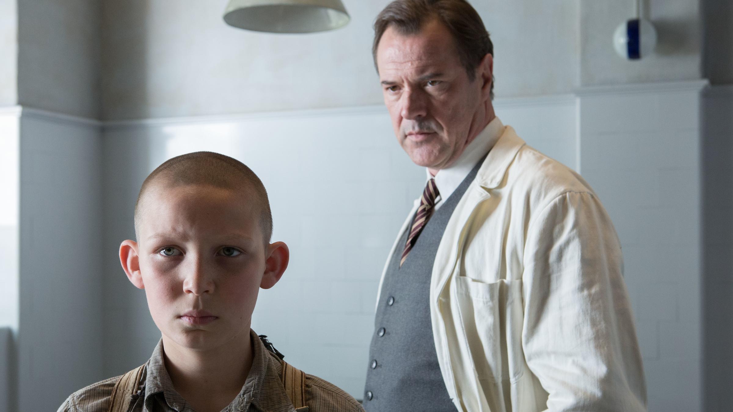 „Nebel im August“: In einem Krankenzimmer steht Dr. Veithausen (Sebastian Koch) hinter dem jungen Ernst (Ivo Pietzcker). Der Doktor betrachtet Ernst eingehend.