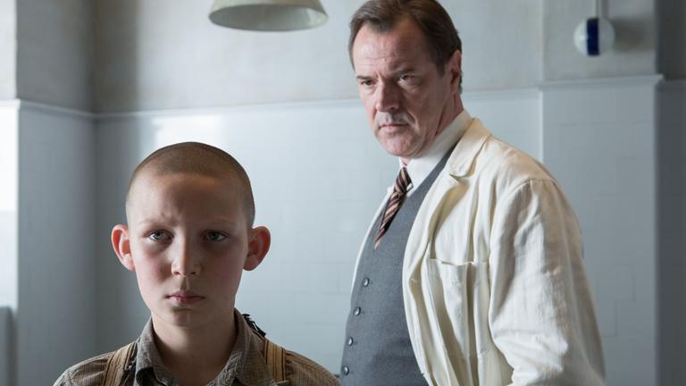 „Nebel im August“: In einem Krankenzimmer steht Dr. Veithausen (Sebastian Koch) hinter dem jungen Ernst (Ivo Pietzcker). Der Doktor betrachtet Ernst eingehend.