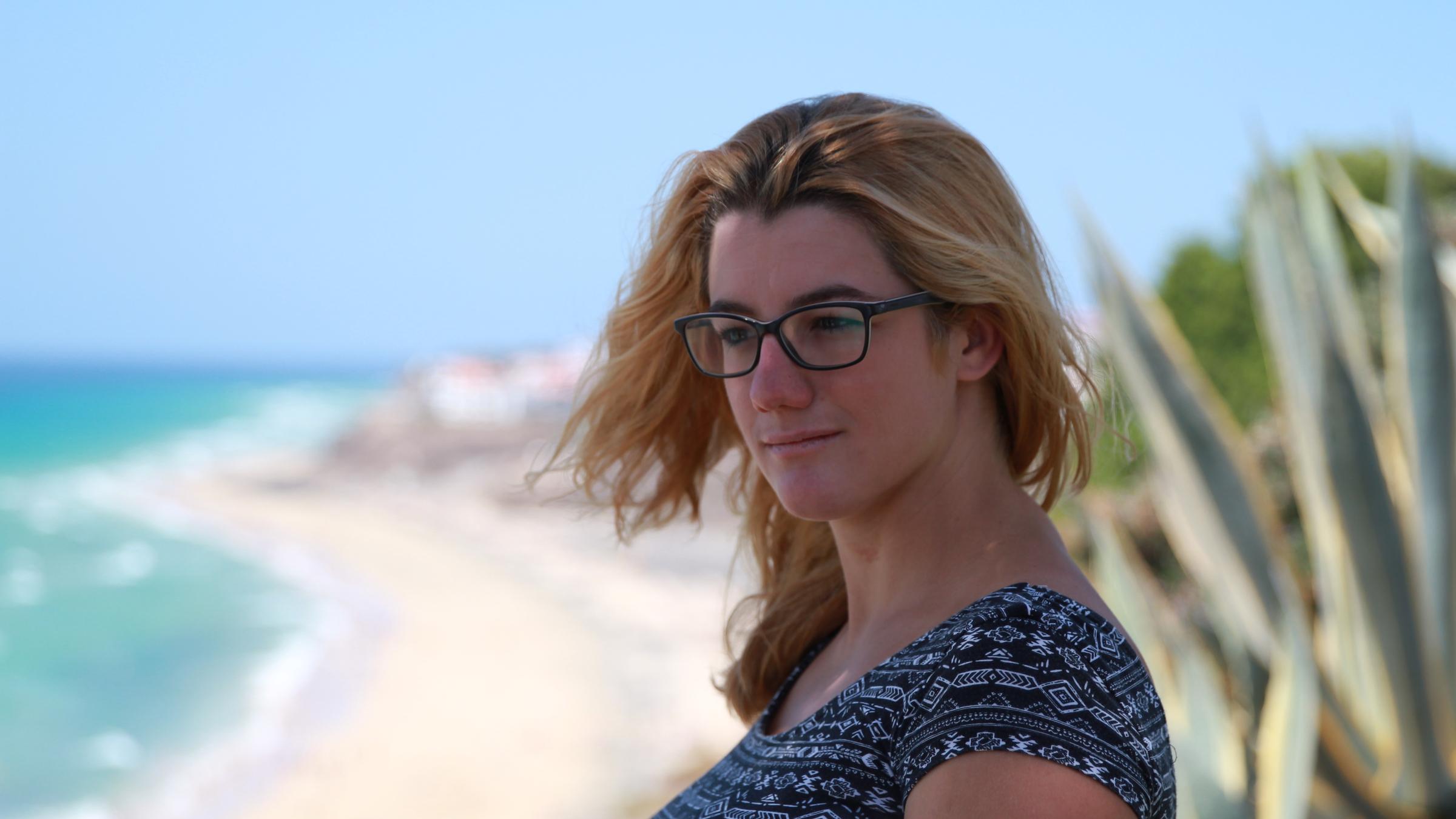 "37°: Ich lebe positiv - Corinne und ihr Schicksal HIV":  Corinne steht an einem gelben Sandstrand. Das Meer ist hellblau und hinter ihr ist eine große Agave zu sehen.