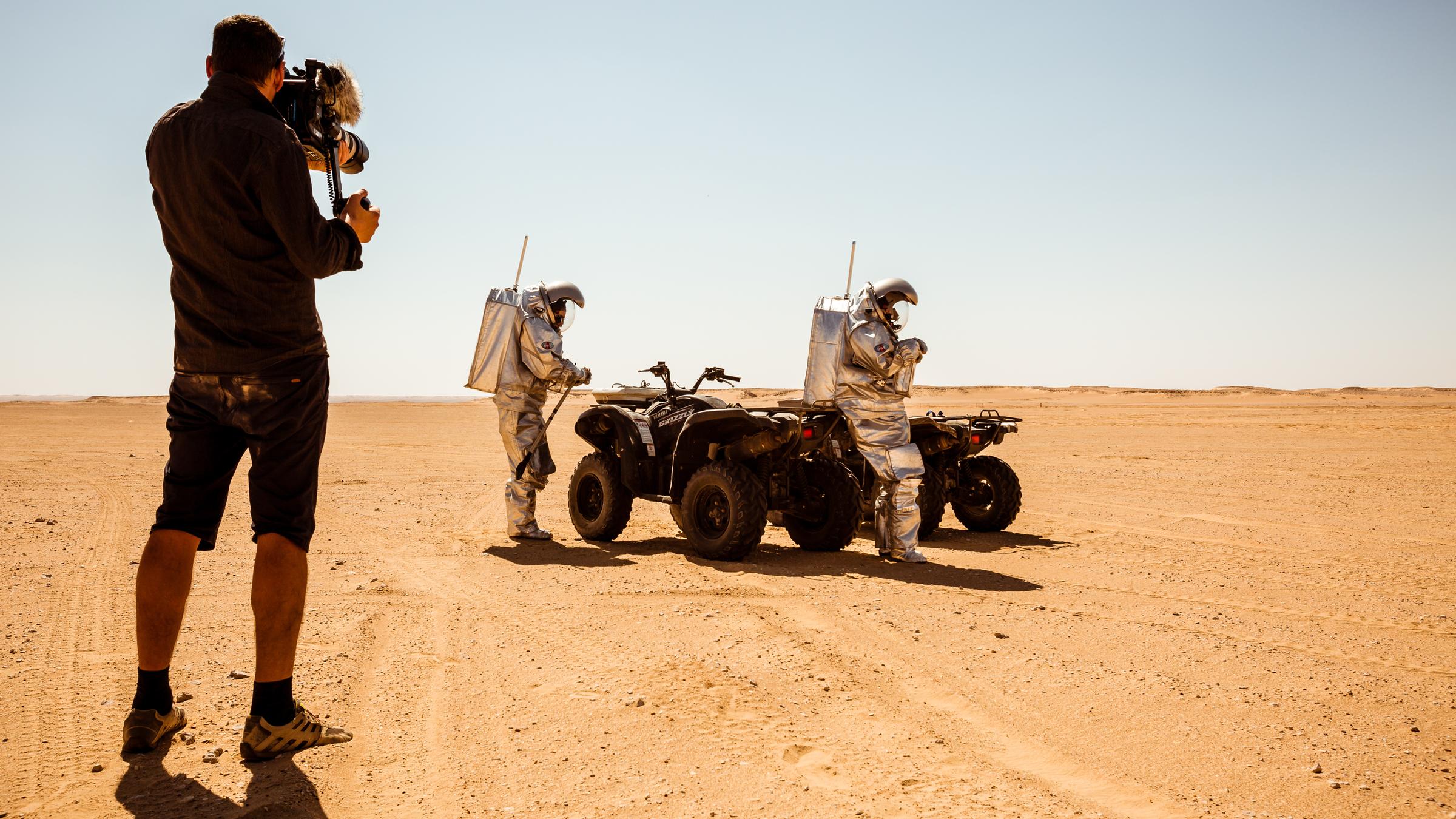 "Terra X: Der Mars - Rätselhafte Wüstenwelt": Wüste im Oman. Ein Mann mit einer Kamera filmt zwei Personen im silbernen Raumanzügen an Motorrädern.