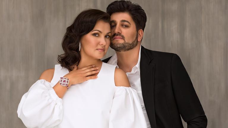 "Anna Netrebko in der Arena di Verona: "Il Trovatore": Die Sopranistin Anna Netrebko trägt ein weißes Oberteil und einen silbernen Armreif mit roten Steinen. Ihr Ehemann, der Tenor Yusif Eyvazov, steht rechts hinter ihr. Er trägt einen schwarzen Anzug und ein weißes Hemd. Die Köpfe der beiden sind eng aneinander.