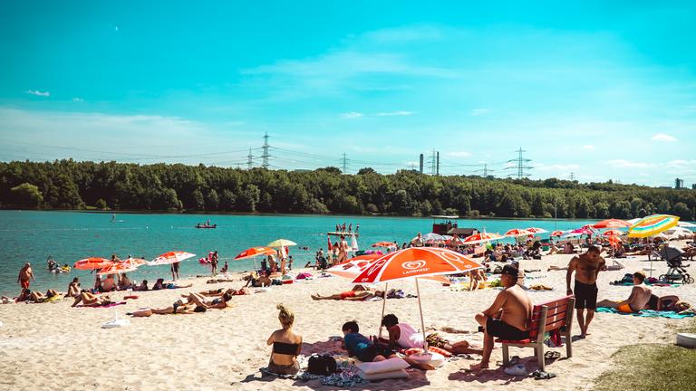 "ZDF.reportage: Schwitzen, schwimmen, feiern - Sommer in der Stadt": Blick über den Strandbereich und den See. Am gegenüberliegenden Ufer des Sees ist liegt ein bewaldeter Bereich. Stromüberlandleitungen ragen über die Bäume. Auf dem Strandabschnitt liegen viele Menschen unter zahlreichen bunten Sonnenschirmen. Im Vordergrund liegen drei Menschen im Sand unter einem orangenen Sonnenschirm. Daneben steht eine rote Bank. Darauf sitzt ein junger Mann in schwarzer Badehose mit schwarzer Kappe. Auf dem See fährt ein Boot und es stehen ein paar Menschen auf der Schwimminsel.