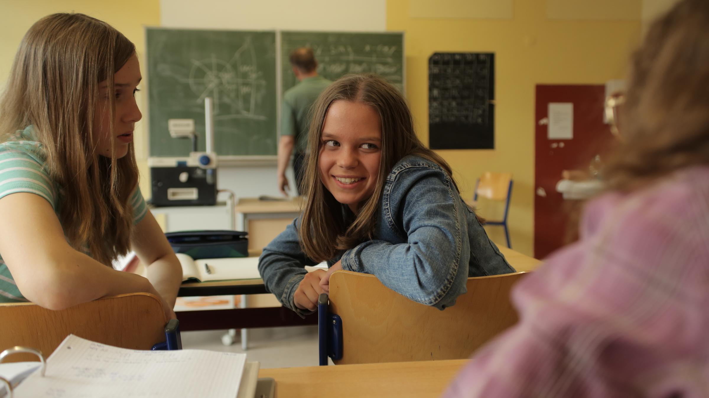 "Aktenzeichen XY... ungelöst - Spezial: Wo ist mein Kind?": Drei jugendliche Mädchen sitzen in einer Schulklasse und unterhalten sich. Der Lehrer schreibt mit dem Rücken zu den Mädchen an die Tafel.
